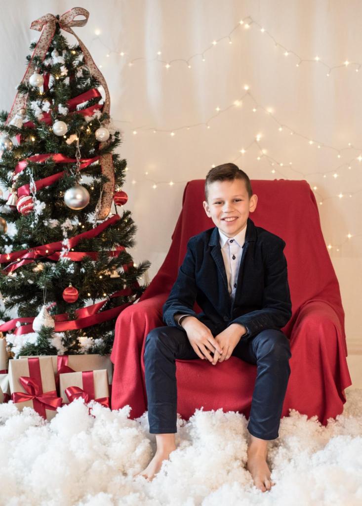Uśmiechnięty chłopiec w granatowym garniturze siedzi boso na białej watolinie obok choinki ozdobionej czerwonymi wstążkami i srebrnymi bombkami, prezenty pod choinką, w tle światełka.