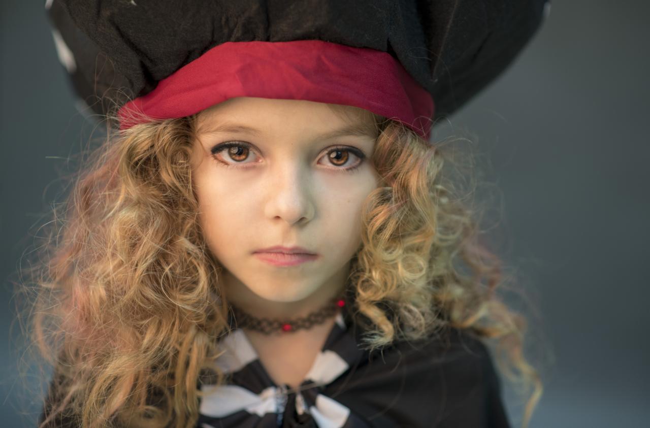 Portret dziewczynki z kręconymi włosami w pirackim kapeluszu i stroju, z delikatnym makijażem oczu.