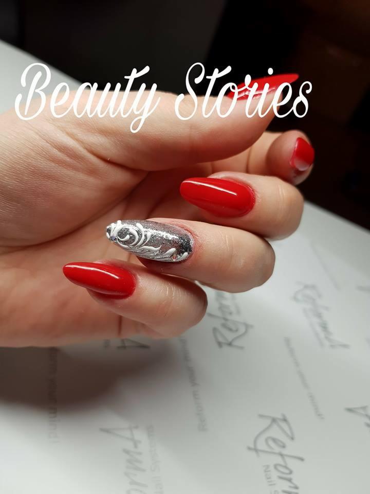 Dłoń z czerwonymi, szpiczastymi paznokciami, jeden z nich ozdobiony srebrnym brokatem i delikatnym, białym wzorem kwiatowym, na tle firmowej podkładki.