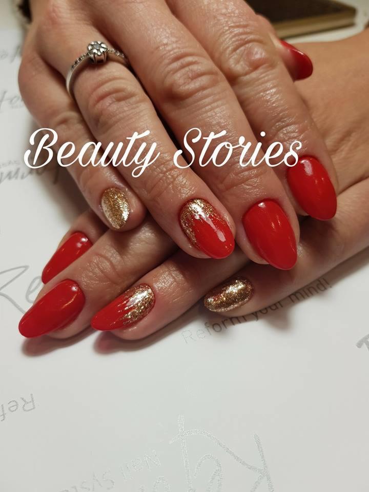 Dłonie z czerwonym manicure hybrydowym i akcentami złotego brokatu na dwóch paznokciach, pierścionek z kwiatowym motywem na palcu wskazującym, logo 'Beauty Stories' nałożone na zdjęcie.