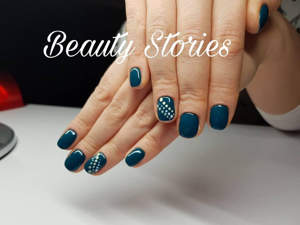 Dłonie z ciemnoturkusowym manicure, jeden paznokieć ozdobiony białymi kropkami, napis 'Beauty Stories' nałożony na zdjęcie.