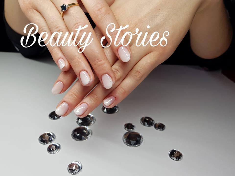 Delikatne ombre na krótkich paznokciach w kształcie migdała, ozdobione drobnymi kryształkami i złotym pierścionkiem z ciemnym oczkiem.