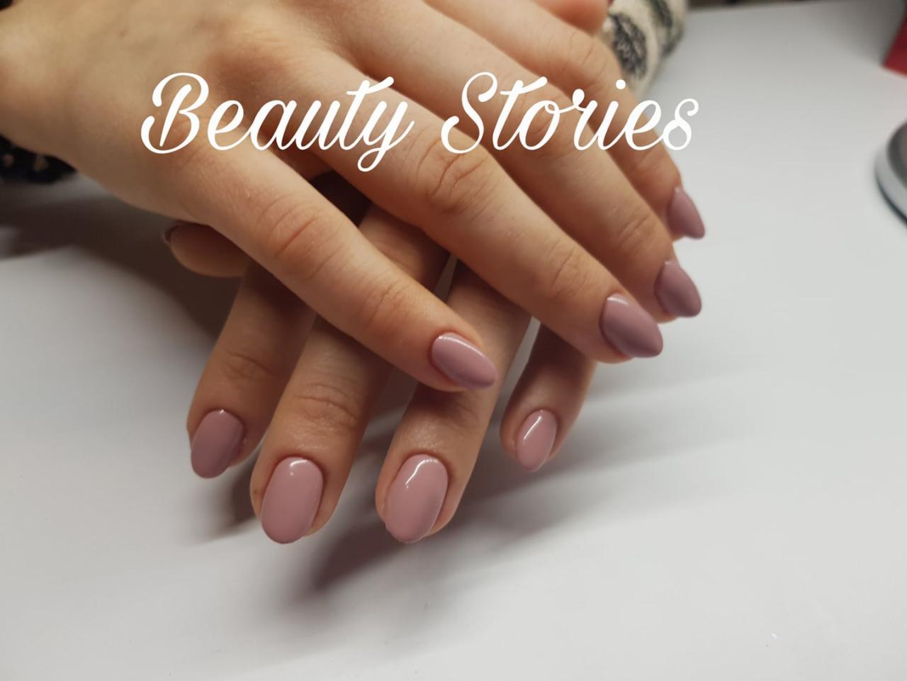 Dłonie z owalnymi paznokciami pomalowanymi na jednolity, pudrowo-różowy kolor, ułożone na białym tle.