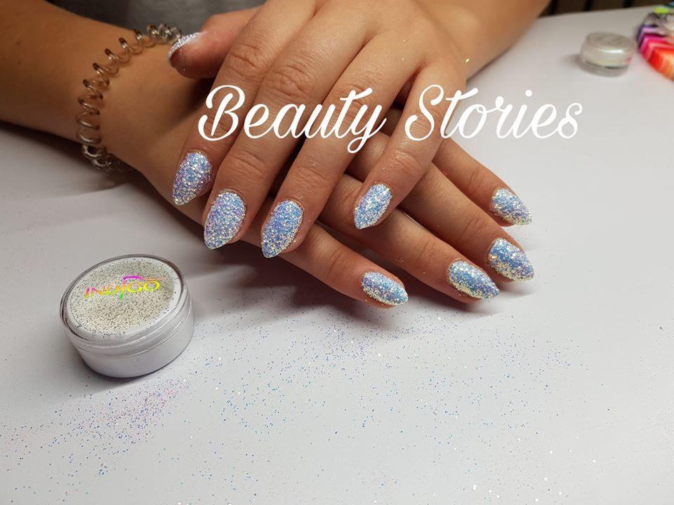 Dłonie z migoczącymi, niebiesko-srebrnymi paznokciami w kształcie migdałów, ułożone na białym blacie, obok słoiczka z brokatem firmy Indigo.