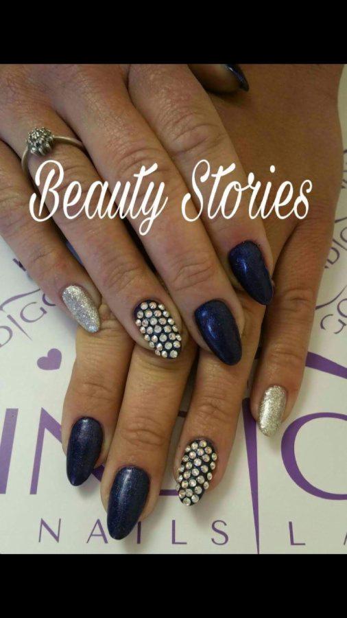 Dłonie z manicure hybrydowym: granatowe i srebrne paznokcie, jeden z paznokci ozdobiony drobnymi kryształkami, pierścionek na palcu wskazującym, logo salonu w tle.