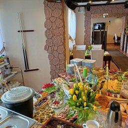 KAERTUR SPÓŁKA Z OGRANICZONĄ ODPOWIEDZIALNOŚCIĄ - Stół szwedzki z bogatym wyborem dań, dekorowany tulipanami i świecami, w restauracji o wystroju z elementami kamienia. Widoczne naczynia, potrawy mięsne, sałatki i pieczywo.