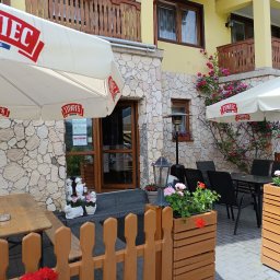 KAERTUR SPÓŁKA Z OGRANICZONĄ ODPOWIEDZIALNOŚCIĄ - Ogródek restauracji z parasolami Żywiec, stolikami i kwiatami w drewnianych donicach. Kamienna elewacja budynku i balkon z drewnianą balustradą w tle.