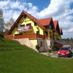 Żółty pensjonat z czerwonym dachem w Nowej Rudzie, z balkonami i kamienną podmurówką, widoczny z przodu na tle zielonego trawnika i błękitnego nieba z chmurami.