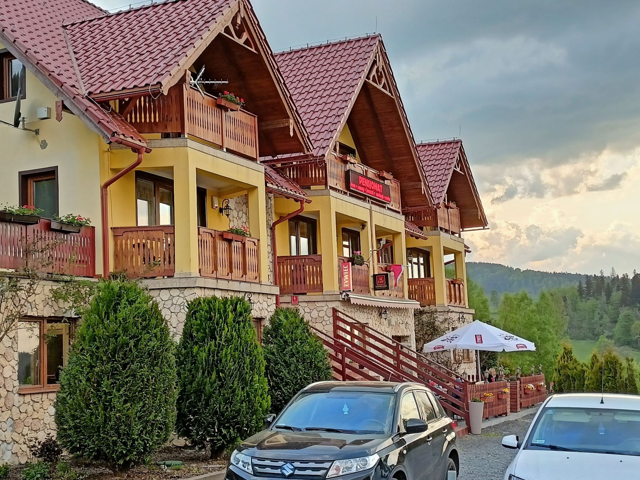 Pensjonat z czerwonym dachem i balkonami, otoczony zielenią. Widoczny fragment parkingu z samochodami. Ujęcie z dołu, podkreślające architekturę budynku.