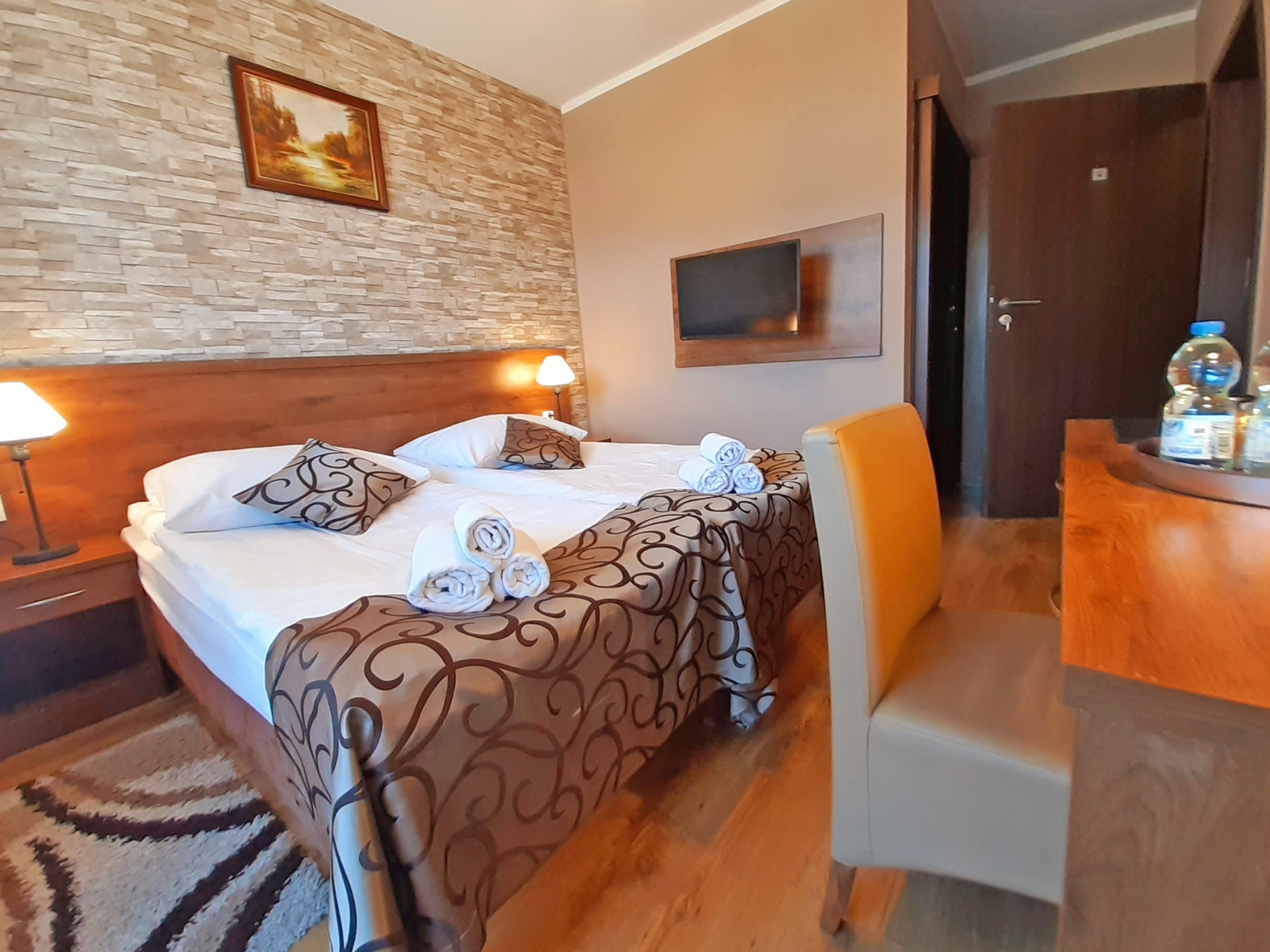 Pokój hotelowy z łóżkiem małżeńskim, telewizorem i stolikiem z wodą mineralną. Ściana z dekoracyjną cegłą i obrazem. Krzesło z żółtym oparciem.