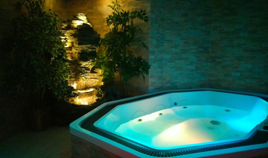 Podświetlany, ośmiokątny jacuzzi z wodą, obok dekoracyjna ściana z kamienia i roślin, tworząca relaksującą atmosferę w spa.