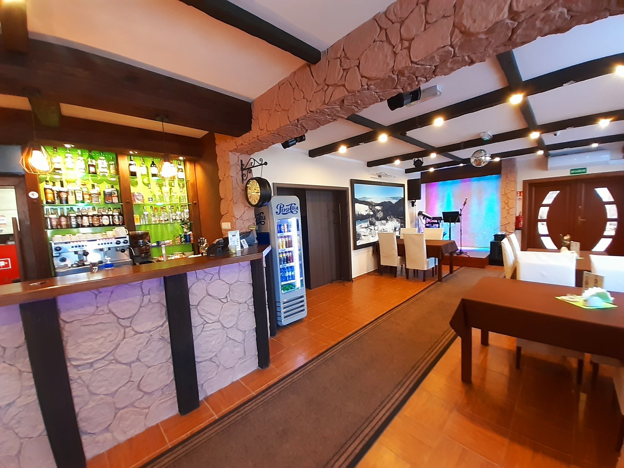 Wnętrze restauracji z barem z kamienną okładziną, stolikami z brązowymi obrusami i sceną z instrumentami w tle. Widoczny dystrybutor Pepsi i dekoracyjne elementy.