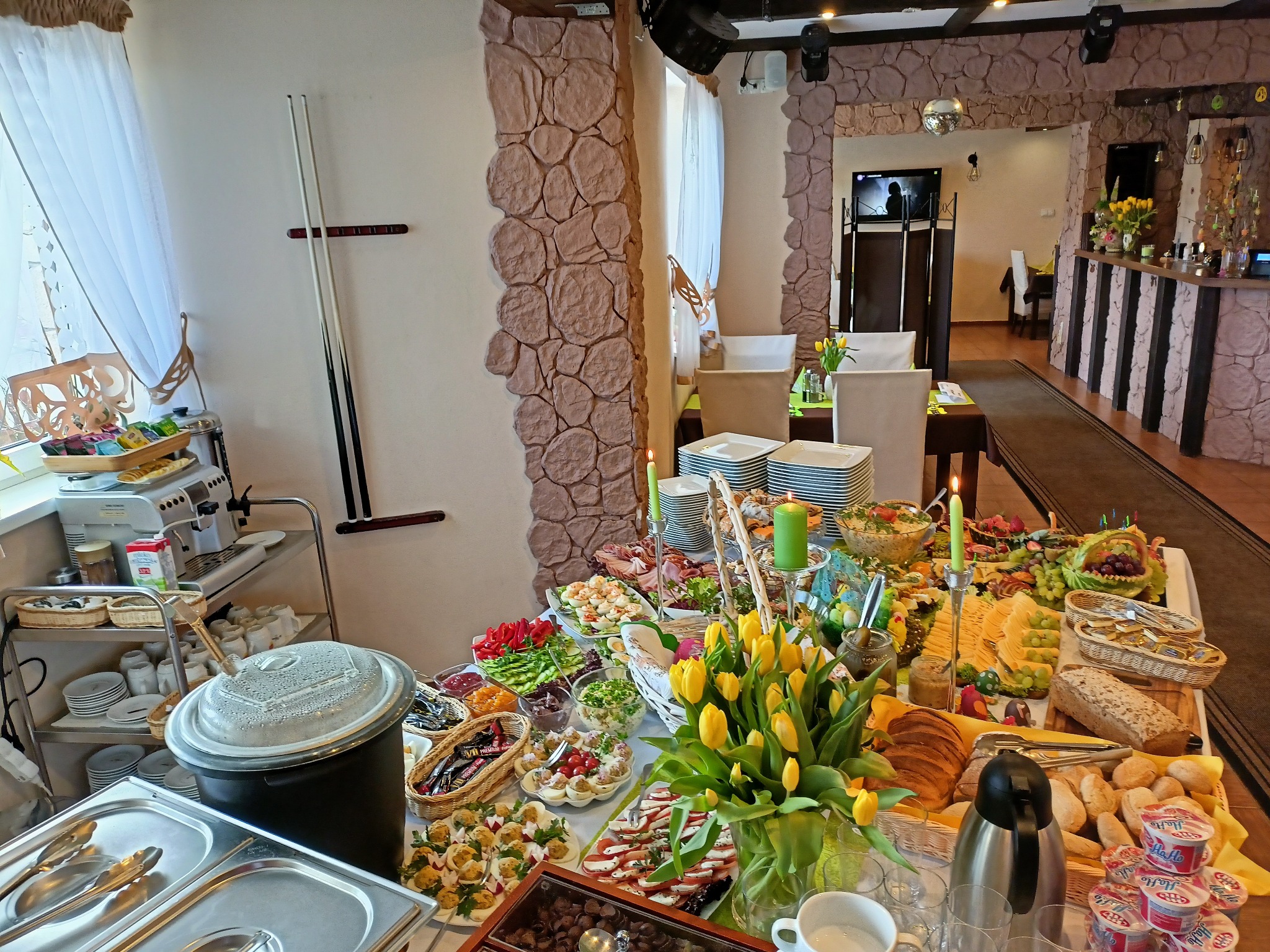 Stół szwedzki z bogatym wyborem dań, dekorowany tulipanami i świecami, w restauracji o wystroju z elementami kamienia. Widoczne naczynia, potrawy mięsne, sałatki i pieczywo.