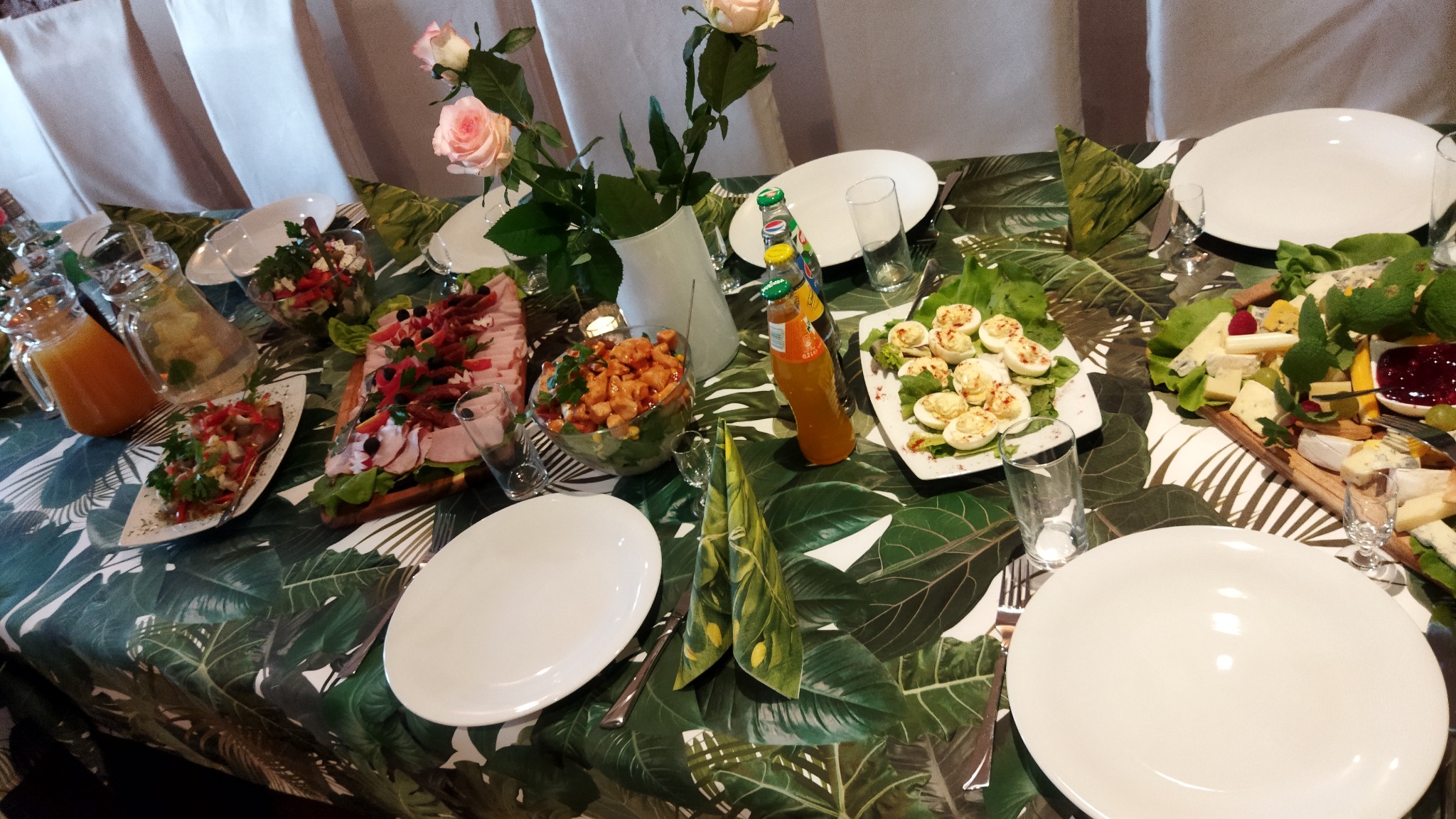 Stół z przekąskami na przyjęciu: sałatki, jajka faszerowane, deska serów, napoje. Dekoracja stołu z motywem liści. Kompozycja kwiatowa z różowych róż.