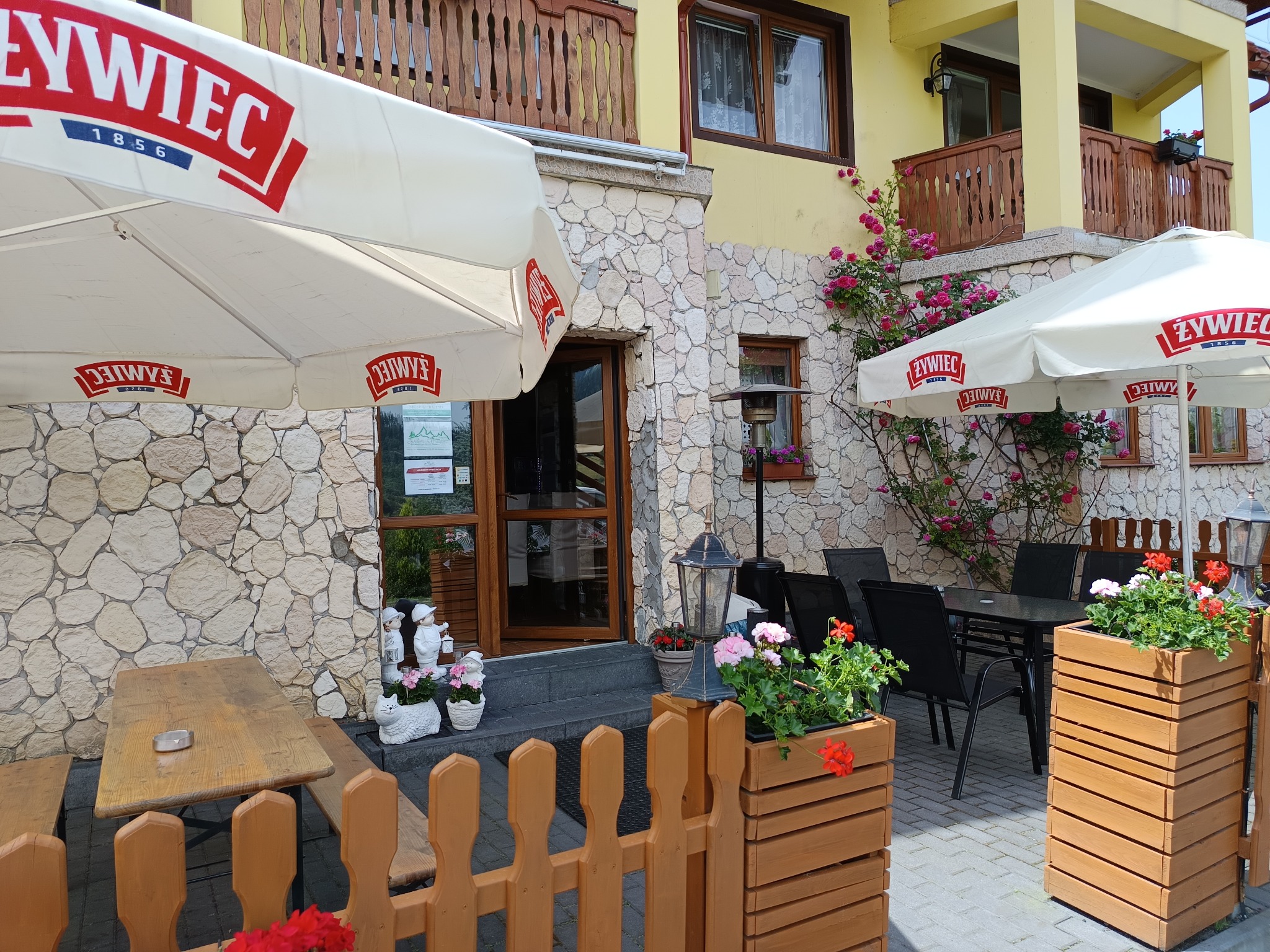Ogródek restauracji z parasolami Żywiec, stolikami i kwiatami w drewnianych donicach. Kamienna elewacja budynku i balkon z drewnianą balustradą w tle.