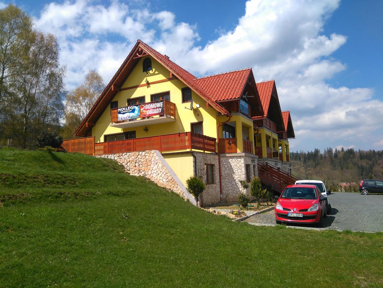 Żółty pensjonat z czerwonym dachem w Nowej Rudzie, z balkonami i kamienną podmurówką, widoczny z przodu na tle zielonego trawnika i błękitnego nieba z chmurami.