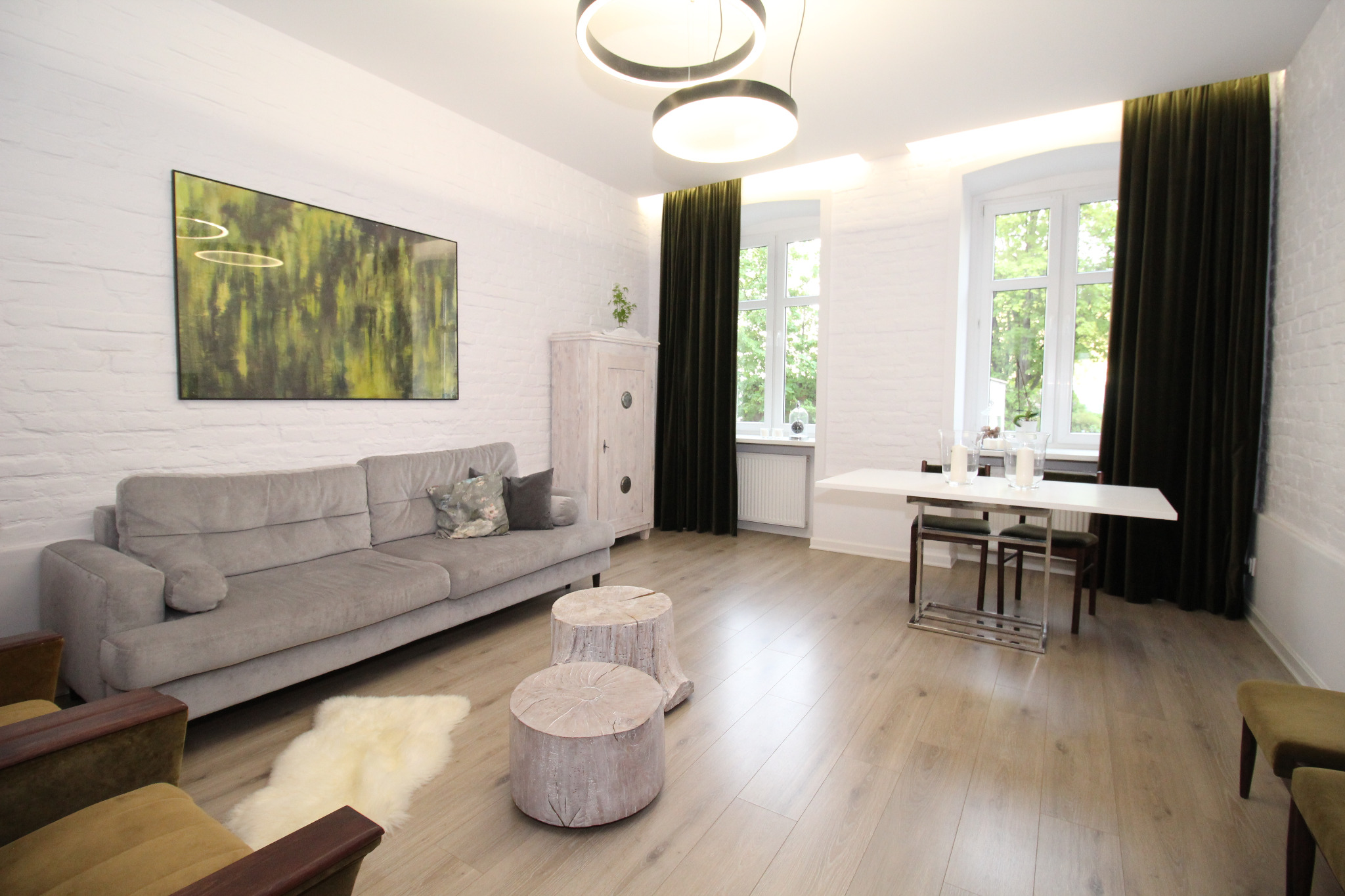 Jasne wnętrze mieszkania z białymi ceglanymi ścianami, szarą sofą, drewnianymi stolikami w kształcie pniaków i obrazem z motywem zieleni na ścianie.