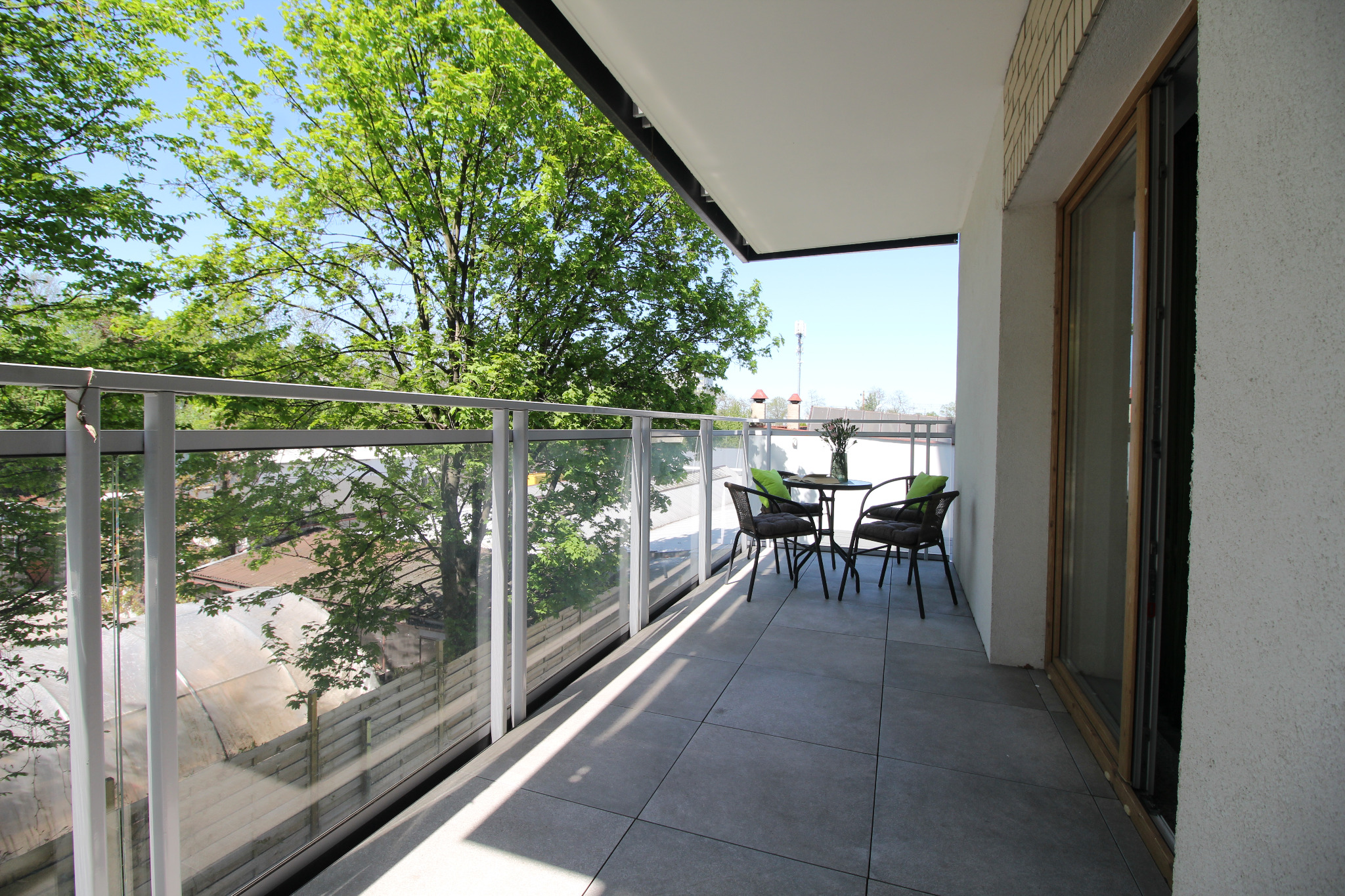 Nowoczesny balkon z szarymi płytkami, szklaną balustradą i zestawem mebli ogrodowych z ciemnymi krzesłami i okrągłym stolikiem z zielonymi poduszkami, widok na zieleń drzew.