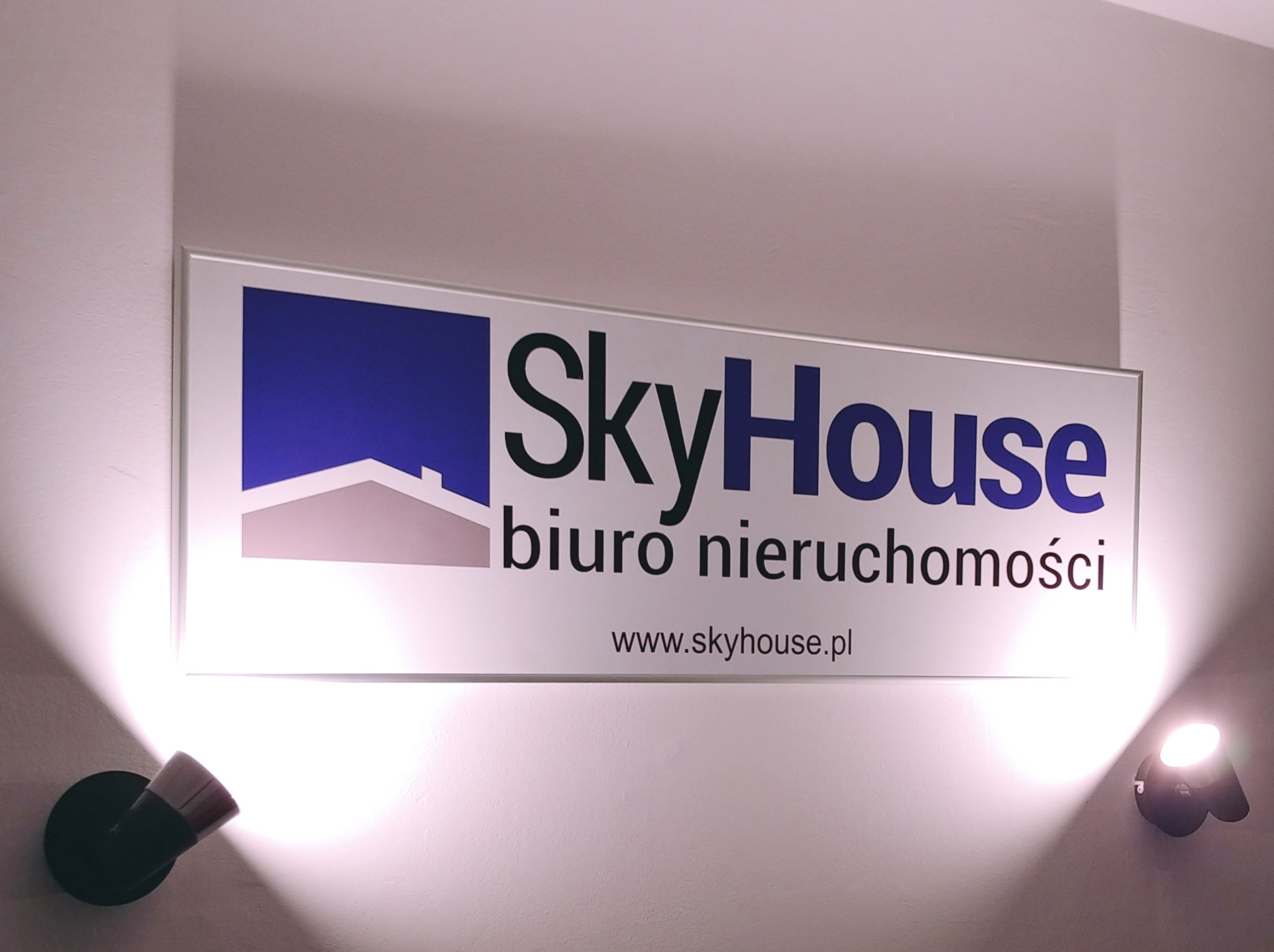 Podświetlany szyld biura nieruchomości SkyHouse z logo i adresem strony internetowej, oświetlony dwoma lampami punktowymi.