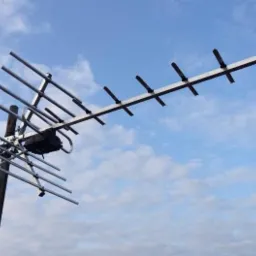 Antena telewizyjna typu Yagi-Uda zamontowana na maszcie, skierowana w stronę błękitnego nieba z delikatnymi obłokami.