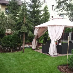 Starannie zagospodarowany ogród z równo przystrzyżonym trawnikiem, licznymi drzewami iglastymi i liściastymi, oraz altaną z zasłonami i meblami ogrodowymi. W tle widoczny budynek mieszkalny.