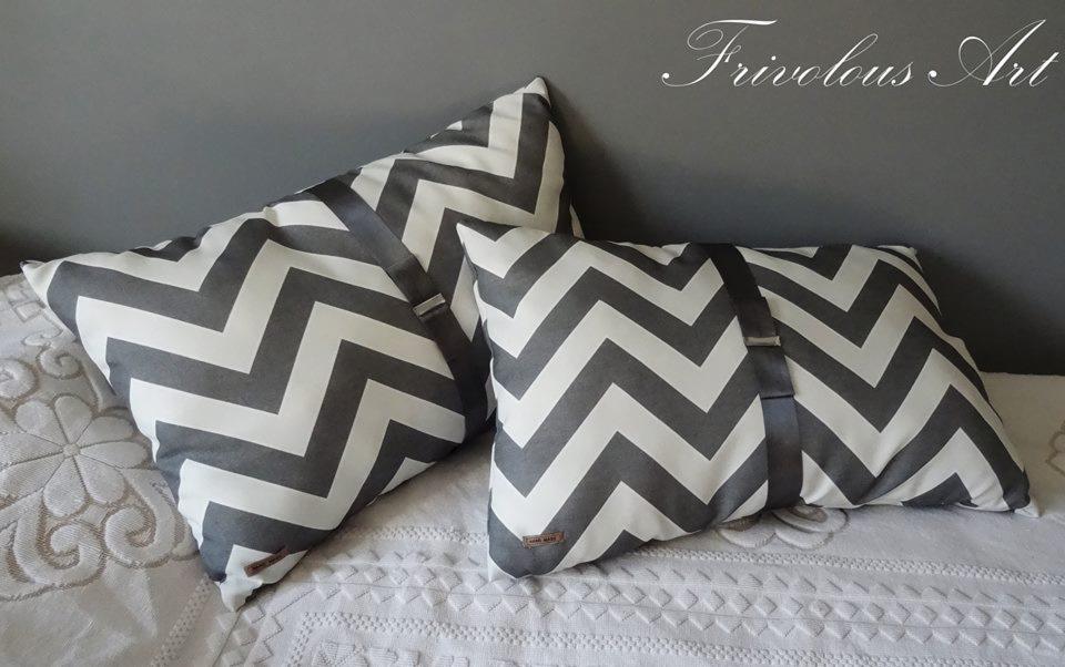Dwie dekoracyjne poduszki w zygzaki szaro-białe, z przymocowanymi pasami, leżą na białej, haftowanej narzucie na łóżko, na tle szarej ściany.