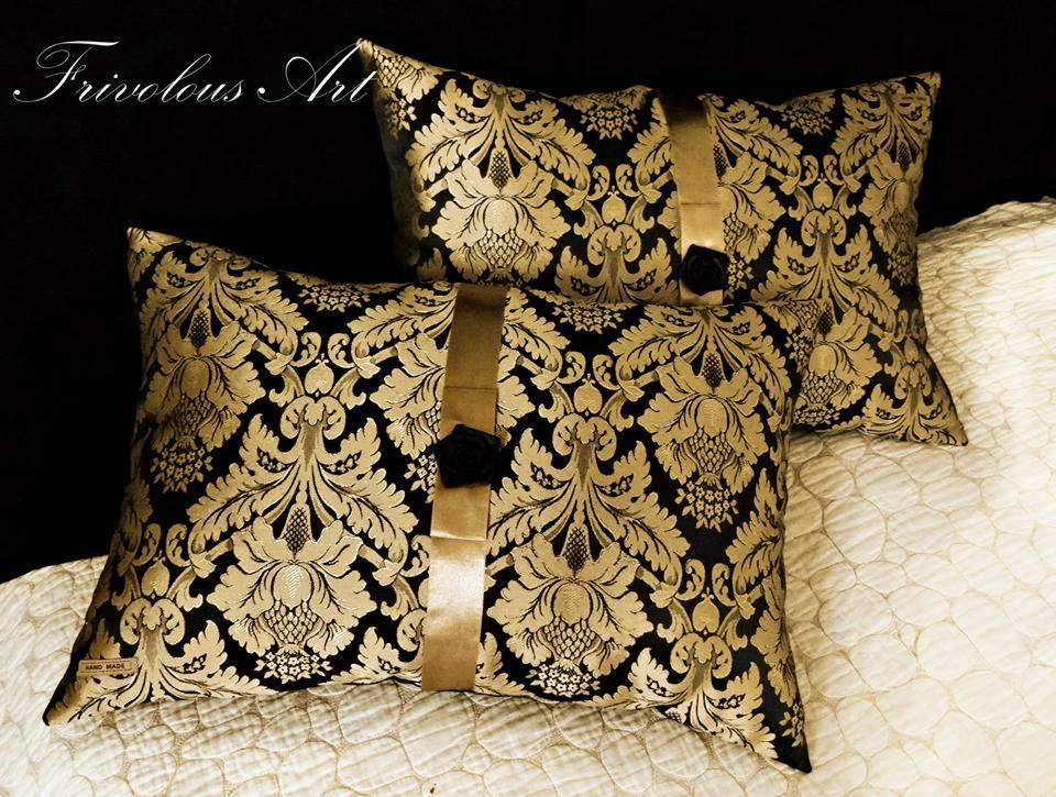 Dwa dekoracyjne poduszki w kolorze czarnym i złotym z barokowym wzorem, przewiązane złotą wstążką z czarną ozdobną różą, leżące na białej pikowanej narzucie.