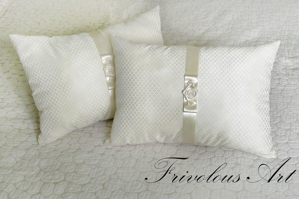 Dwie dekoracyjne, kwadratowe poduszki w odcieniach bieli z subtelnym wzorem, ozdobione satynowymi wstążkami i aplikacjami w kształcie różyczek, leżące na pikowanej narzucie.