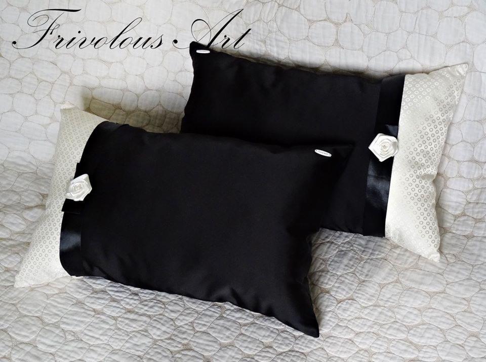 Dwie dekoracyjne poduszki: jedna czarna, druga z czarno-białym wzorem, obie z satynową wstążką i ozdobną różą, ułożone na pikowanej narzucie.