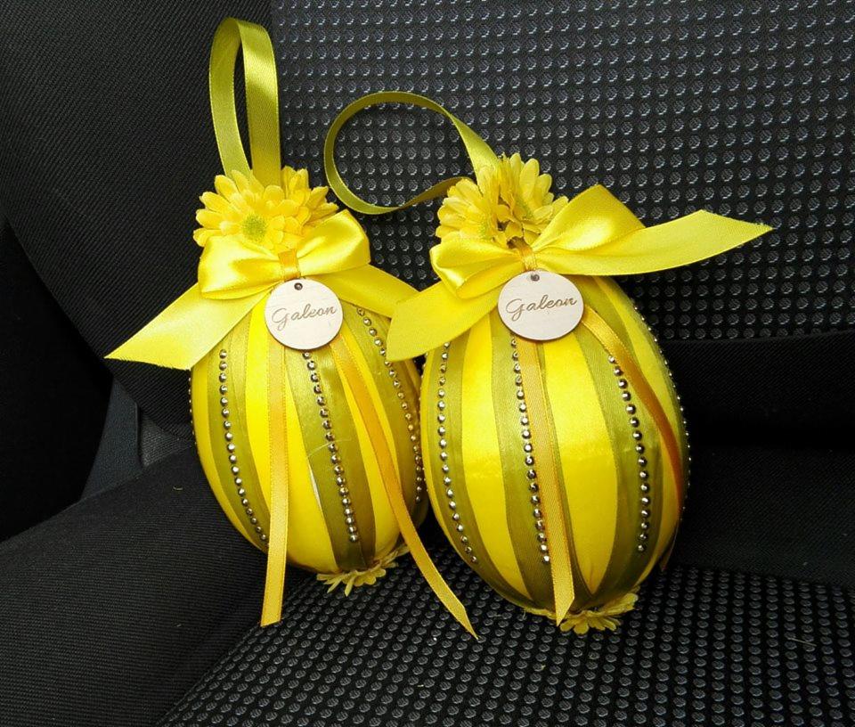 Dwa żółte, ozdobne jajka z kokardami i zawieszkami z napisem 'Galeon', umieszczone na ciemnym, teksturowanym tle.