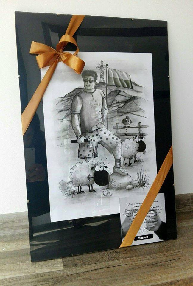 Oprawiony w czarną ramę rysunek przedstawiający mężczyznę w okularach, z siekierą w ręku, stojącego obok trzech owiec na tle krajobrazu z wiatrakiem i polską flagą, całość ozdobiona złotą wstążką.