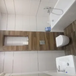 Wykończona łazienka z białymi płytkami ściennymi i drewnopodobnymi płytkami podłogowymi, z wnęką w ścianie wyłożoną drewnem, zawierającą rolkę papieru toaletowego, oraz białą armaturą: sedesem...