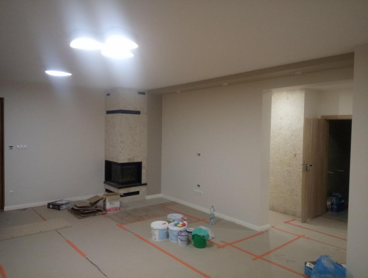 Wnętrze pokoju w trakcie prac wykończeniowych; widoczne pomalowane na biało ściany, obudowany kamieniem kominek, zabezpieczona folią podłoga z widocznymi fragmentami parkietu, puszki z farbą...