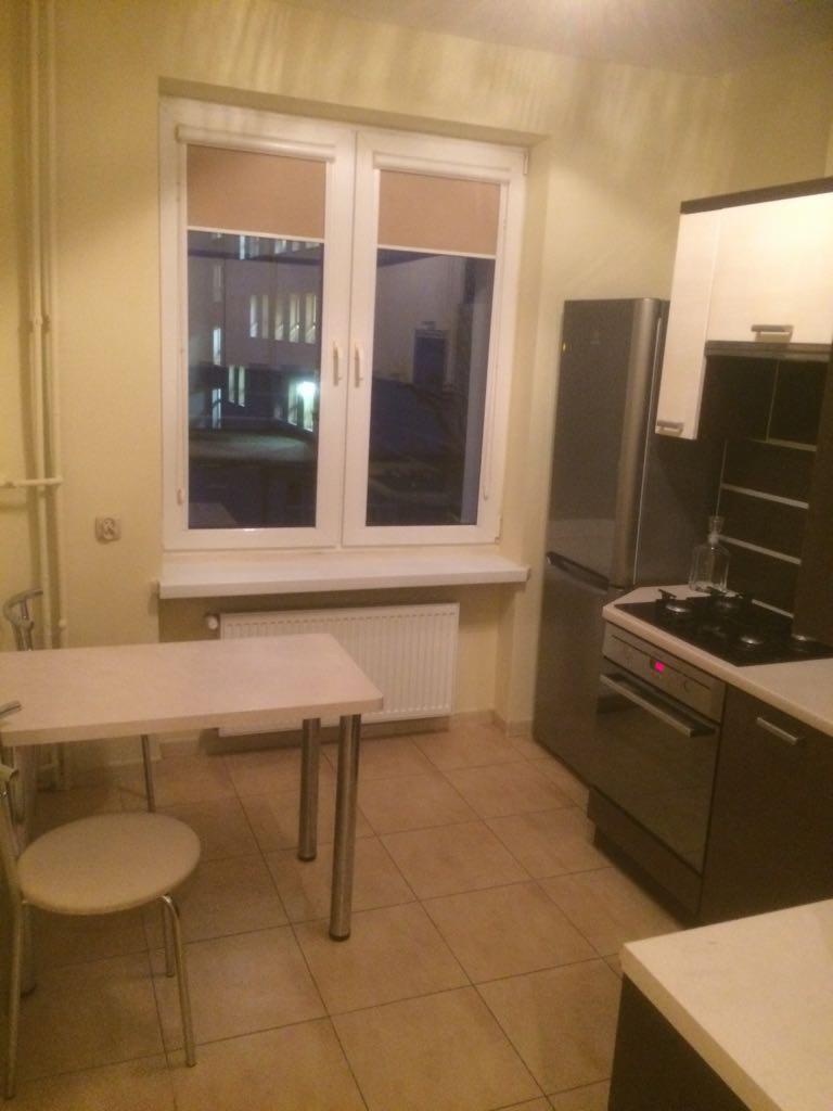 Kuchnia z beżowymi ścianami i podłogą, widoczne okno z roletami, stół z krzesłem, lodówka i kuchenka, oświetlenie nocne za oknem.