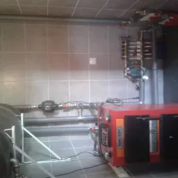 Czerwony piec CO RAKOCZY POPTERG z podłączonymi rurami i instalacją grzewczą w kotłowni wyłożonej szarymi płytkami.