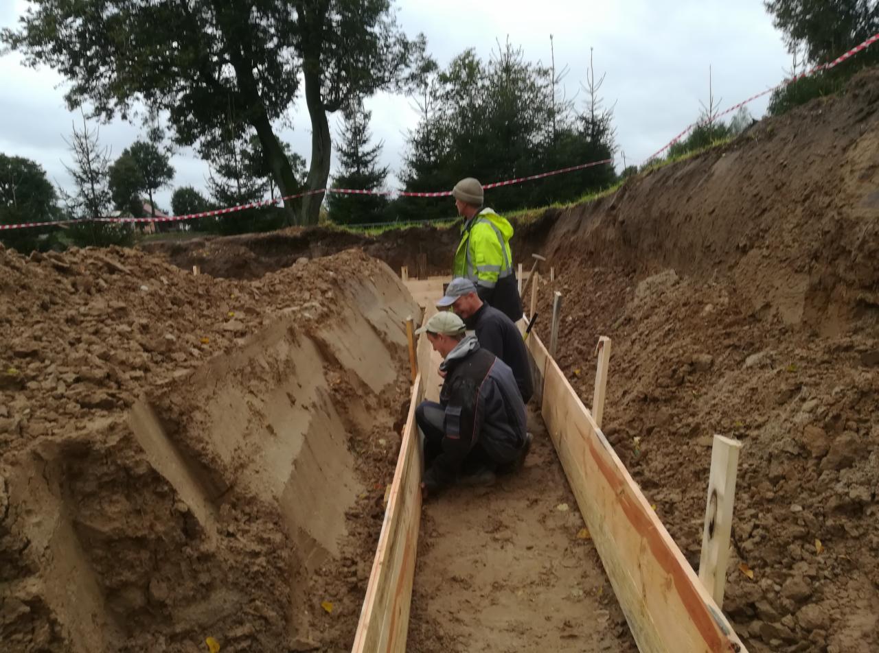 Trzech mężczyzn w odzieży roboczej przygotowuje szalunek w wykopie ziemnym pod fundament budynku, widoczne deski i ziemia.