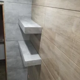 Dwie półki z płytek ceramicznych zamontowane na ścianie wyłożonej płytkami imitującymi drewno i szare płytki w łazience.