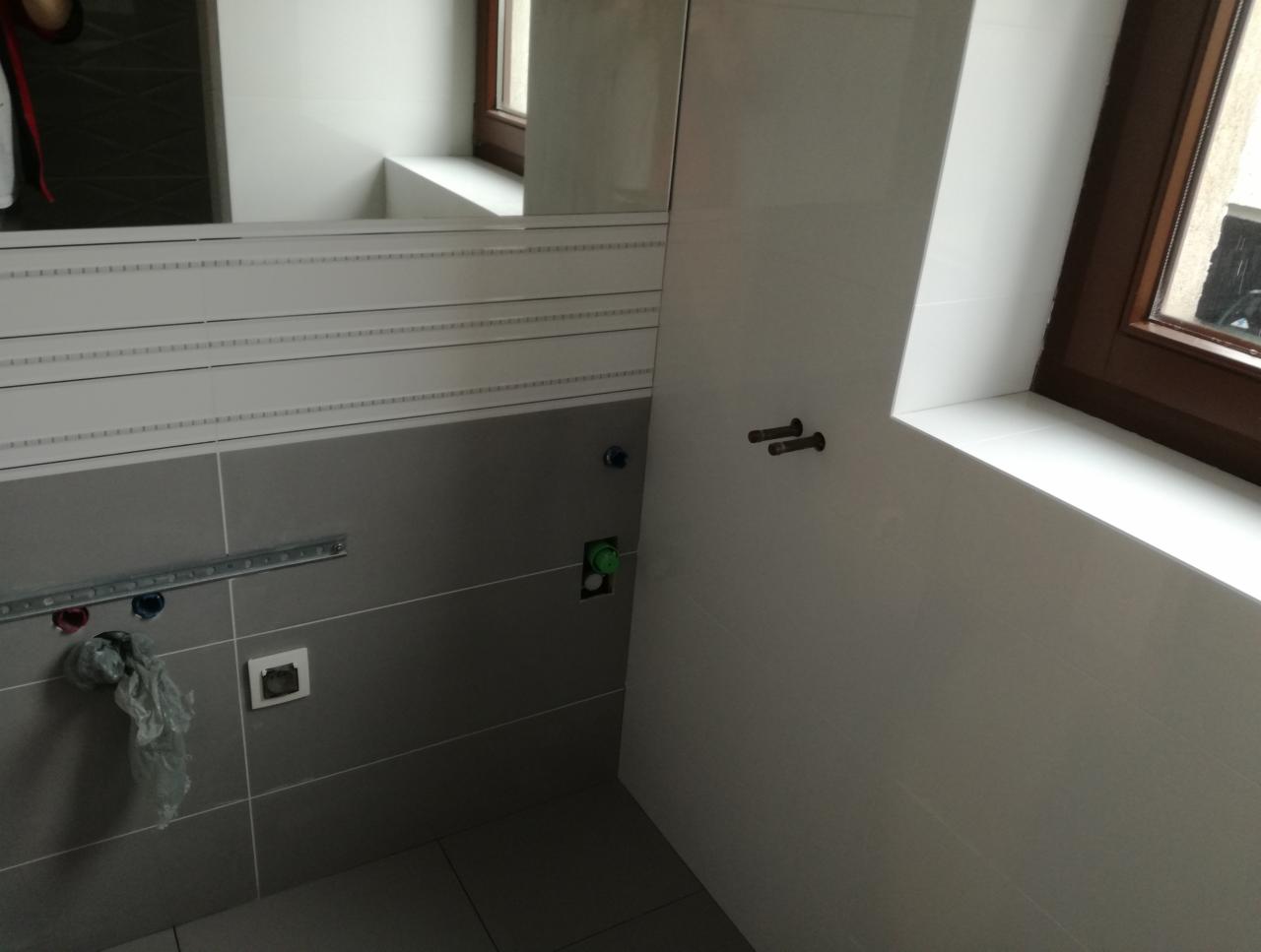 Szara ściana w łazience z częściowo ułożonymi białymi płytkami, widoczne instalacje wodno-kanalizacyjne i odblask lustra.