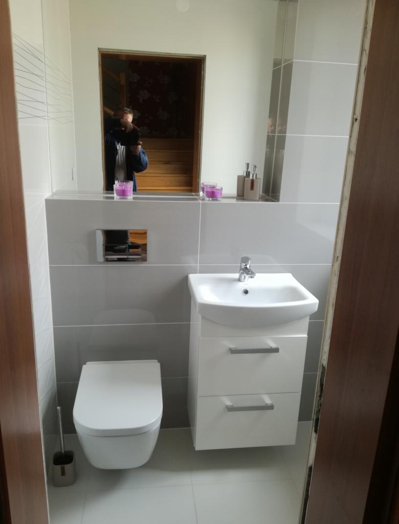 Nowoczesna łazienka z podwieszaną toaletą, umywalką z szafką i szarymi płytkami, widoczne akcesoria łazienkowe i odbicie osoby w lustrze.