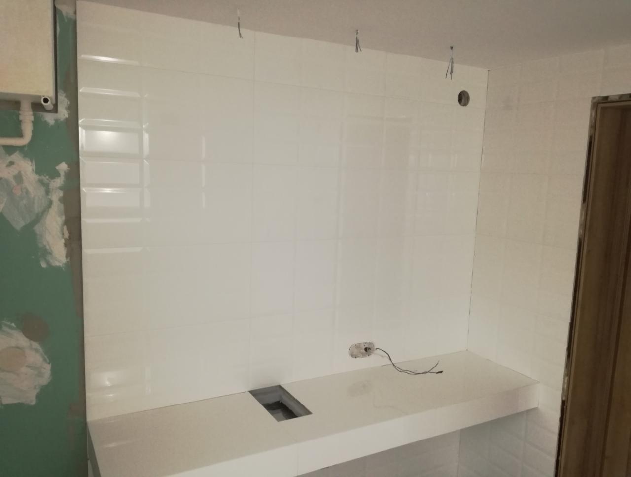 Ściana łazienki wyłożona białymi, błyszczącymi płytkami, z widocznymi śladami prac wykończeniowych na ścianie po lewej stronie oraz wystającymi przewodami elektrycznymi i otworem na gniazdko.