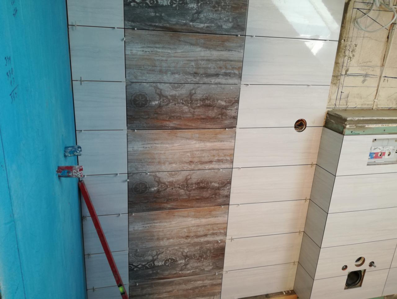 Ściana w trakcie wykańczania z ułożonymi płytkami w odcieniach bieli i brązu, widoczne krzyżyki dystansowe i poziomica przy niebieskiej płycie kartonowo-gipsowej.