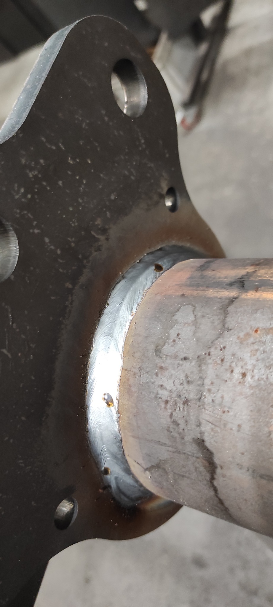 Połączenie spawane rury z metalowym elementem z otworami montażowymi, widoczne ślady po spawaniu i niedoskonałości spoiny.