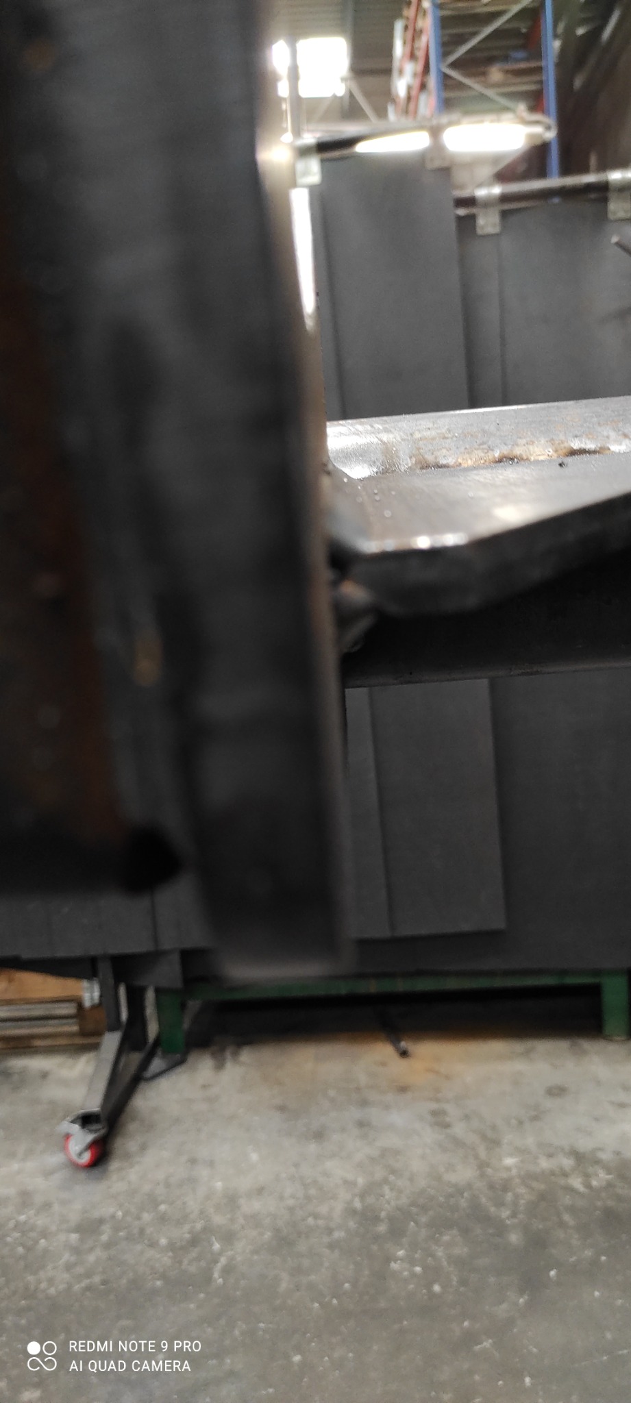 Spoina łącząca dwie metalowe płyty, widoczna z bliska, na tle stanowiska spawalniczego z czarnymi osłonami.