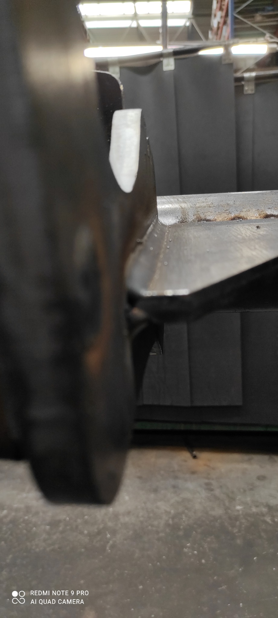 Zbliżenie na spoinę na metalowej konstrukcji, widoczne ślady po obróbce i drobne nierówności, tło stanowią ciemne, pionowe panele.