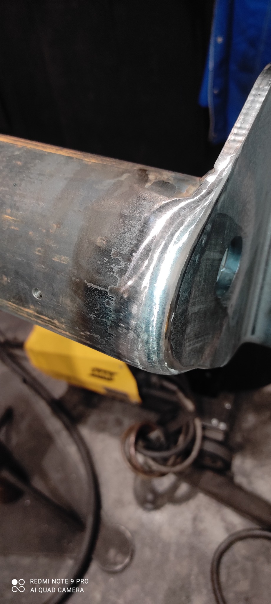 Spoina na metalowym elemencie konstrukcyjnym, widoczne ślady obróbki i żółta spawarka w tle.