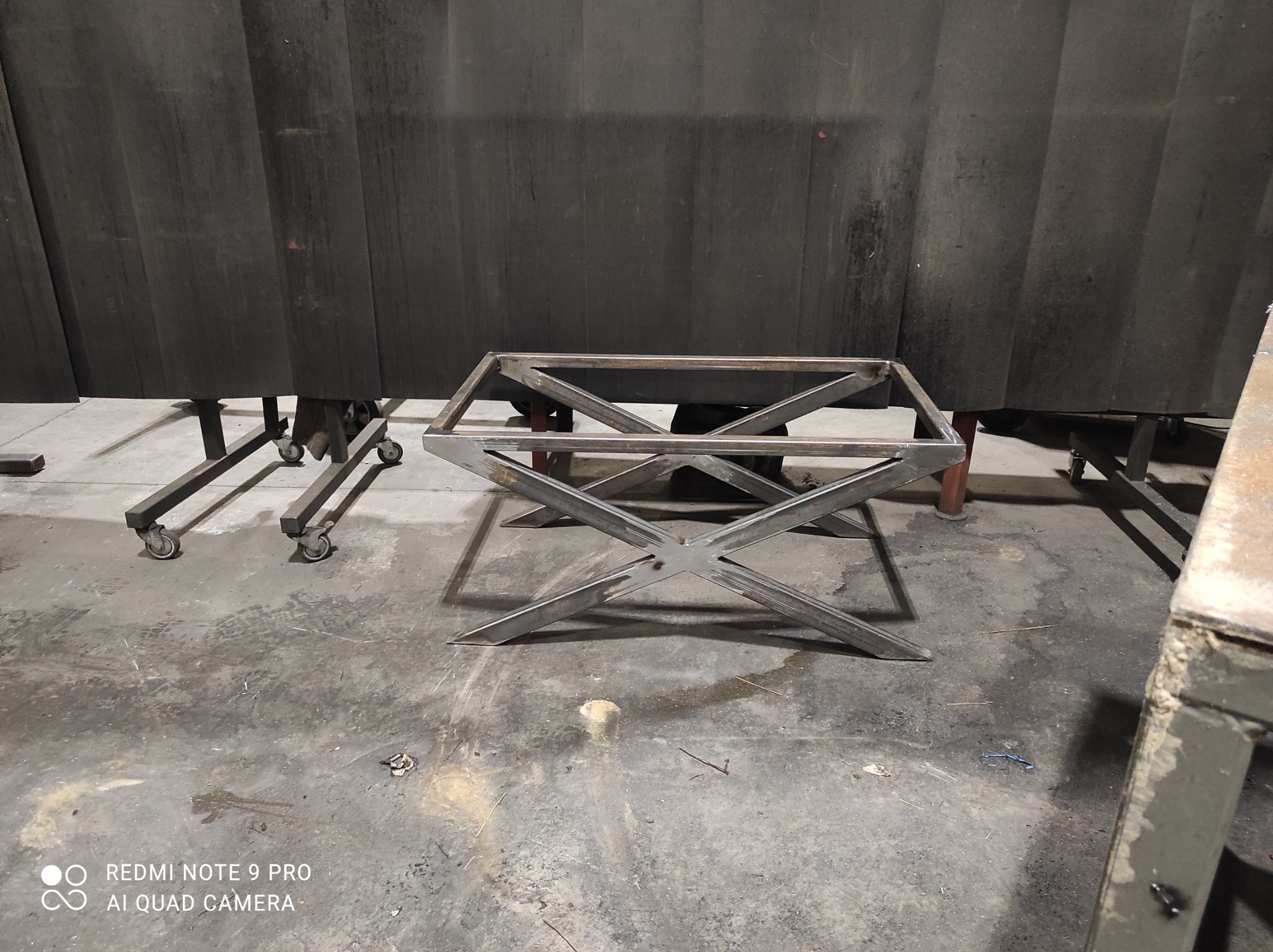 Metalowa konstrukcja stolika kawowego z nogami w kształcie litery X, spawana w warsztacie, na tle zasłon spawalniczych.