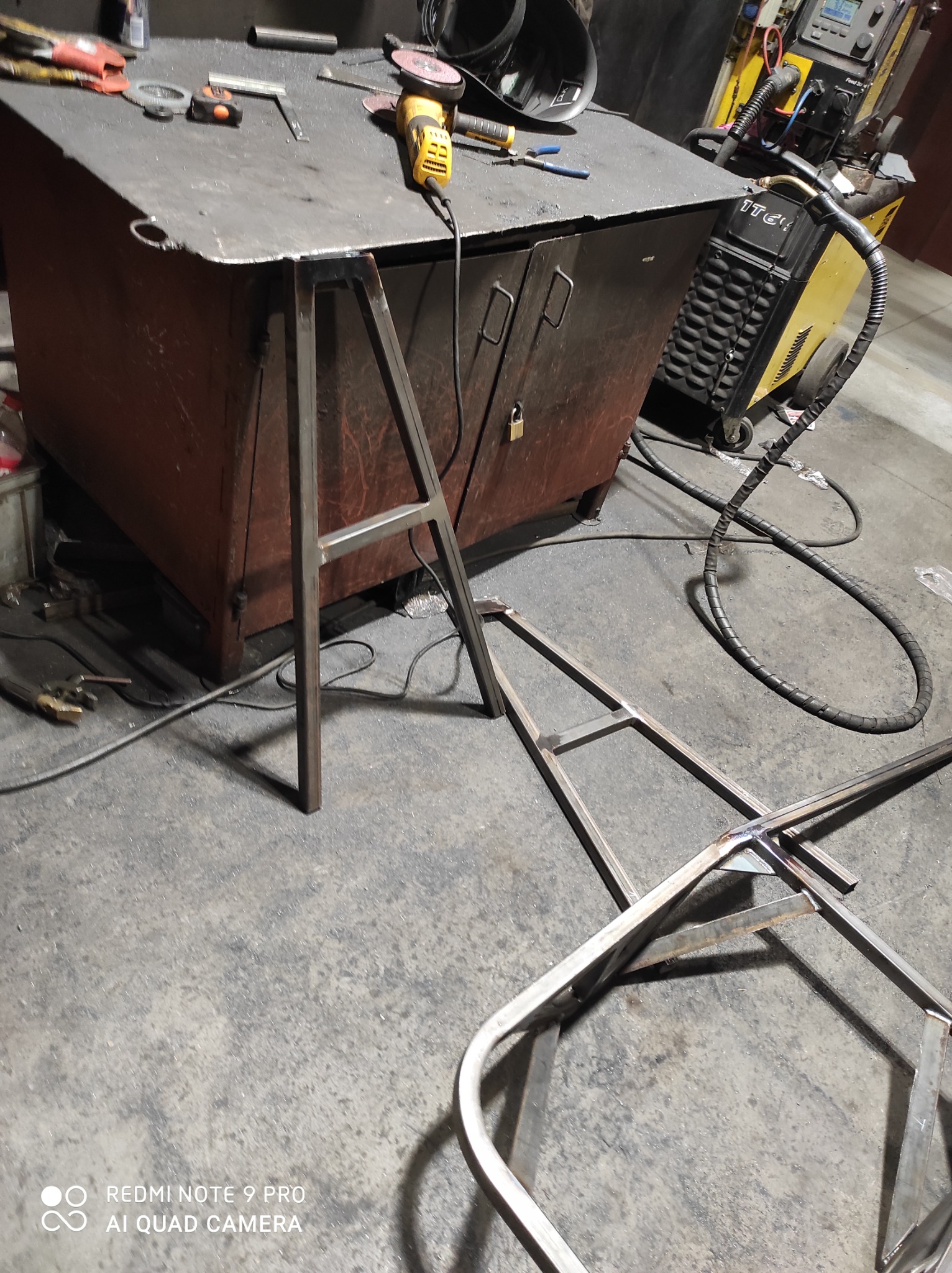 Metalowe elementy konstrukcyjne spawane na warsztatowym stole, widoczna spawarka, szlifierka kątowa i narzędzia.