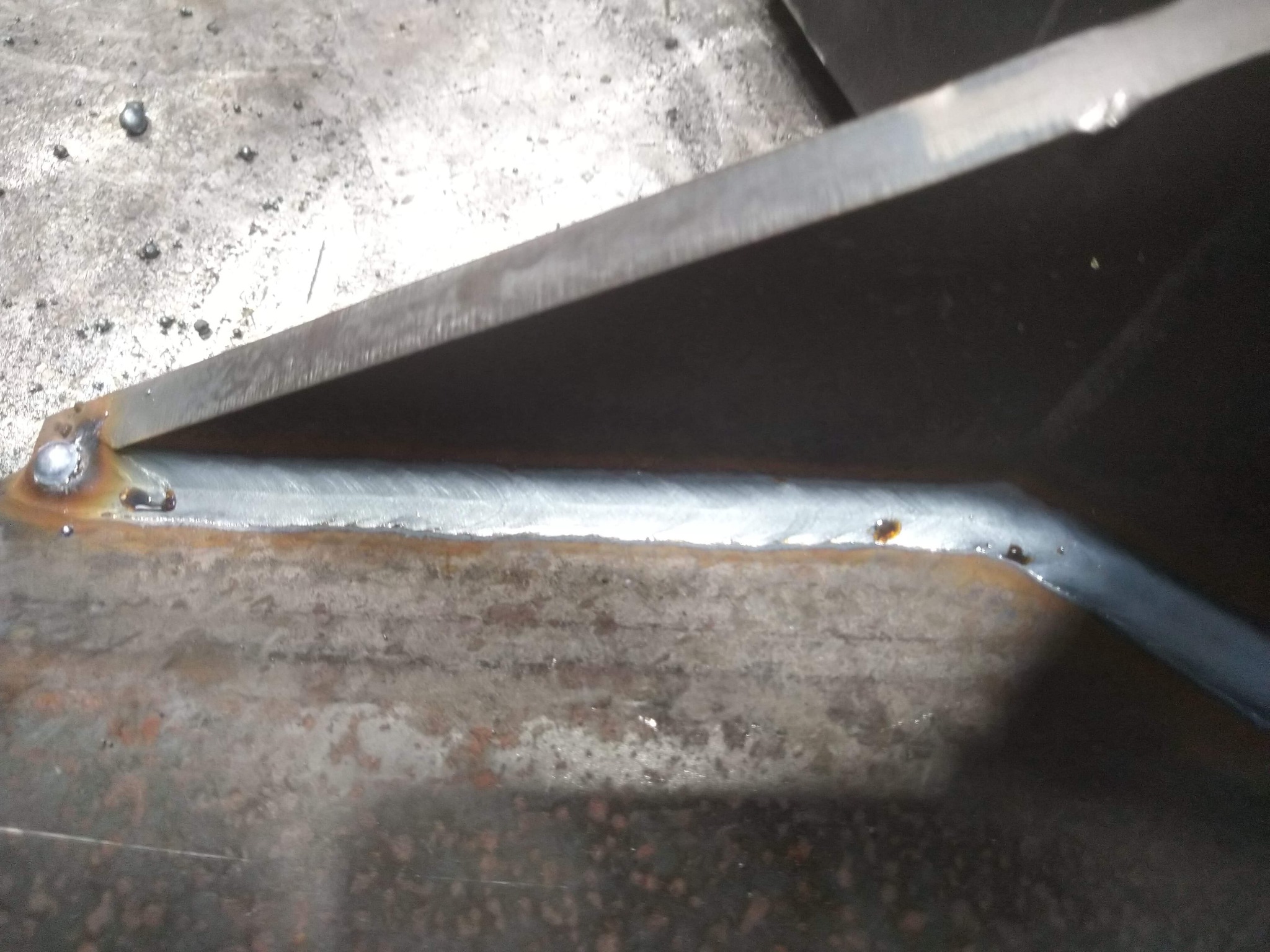 Połączenie spawane dwóch metalowych elementów pod kątem, widoczny spaw na szorstkiej powierzchni roboczej.