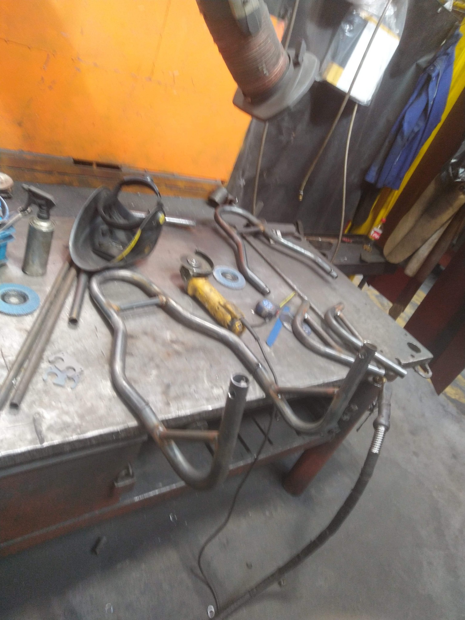 Metalowa konstrukcja w trakcie obróbki na warsztatowym stole, widoczna maska ochronna, szlifierka kątowa i inne narzędzia.