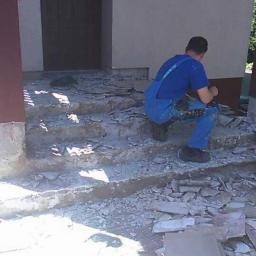 Rem-Bud Bartosz Żemła - Mężczyzna w niebieskim kombinezonie skuwa stare płytki z betonowych schodów wejściowych do domu, przygotowując podłoże pod nowe wykończenie, z boku leżą stosy zdjętych płytek.